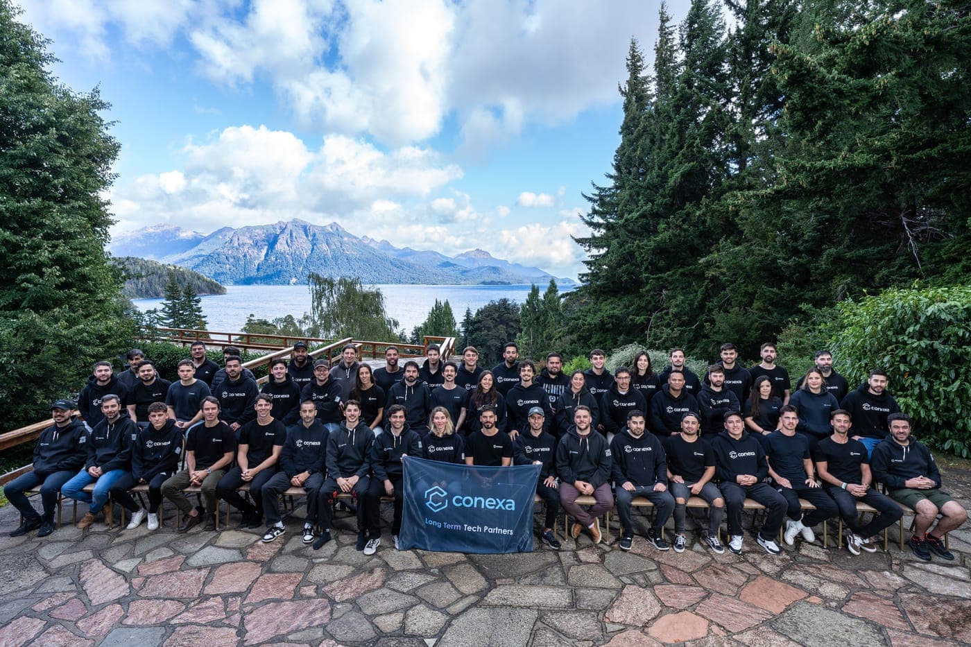 Large group photo of Conexa employees seated and standing in rows on a stone terrace, wearing matching black Conexa t-shirts, with a "Conexa – Latin America Tech Partners" banner in front, and a mountain lake framed by tall pine trees in the background.