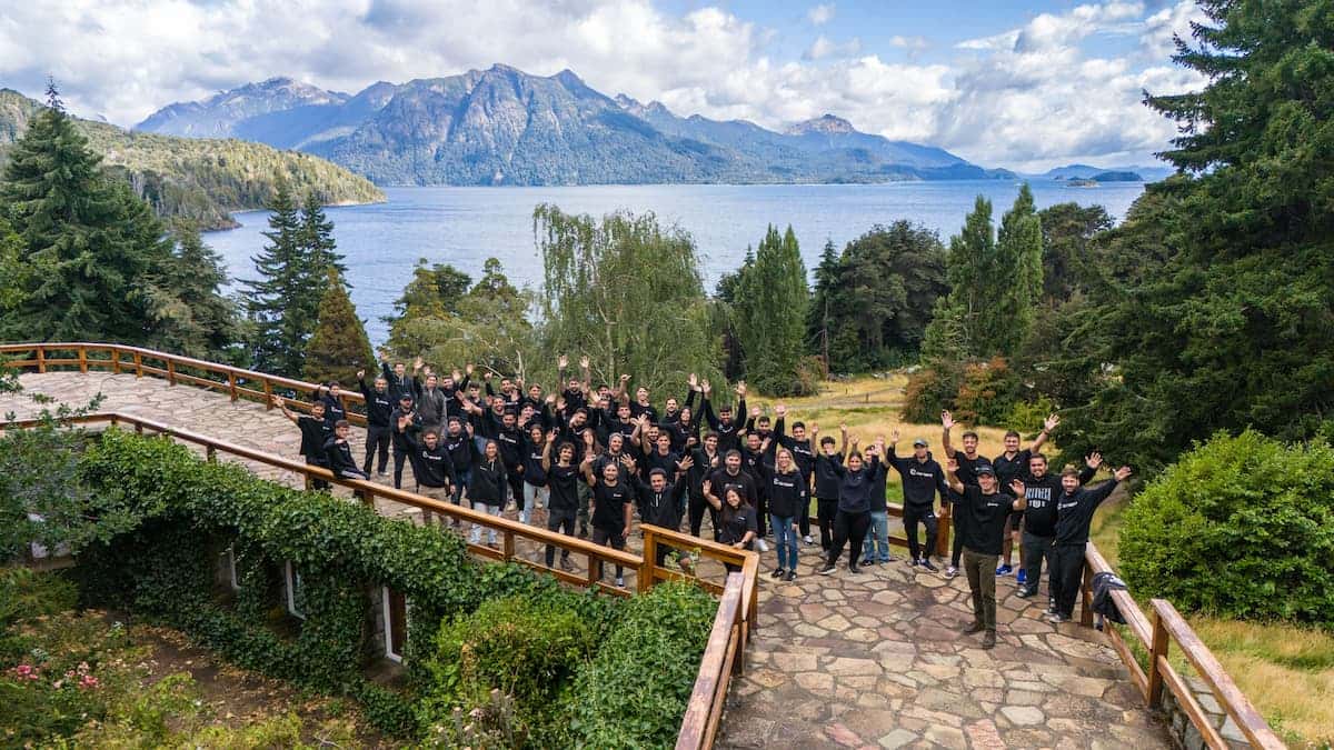 Group of Conexa employees standing on a stone terrace overlooking a mountain lake, cheering with raised arms, surrounded by forested hills under a partly cloudy sky.